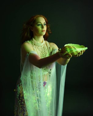 Peri masalı kostümü giymiş kızıl saçlı bir deniz kızı perisinin fantezi stüdyosu portresi. Klasik poz, deniz kabuğunu havada süzülen kumaş ve ruhsal kasvetli stüdyo ışıklandırmasıyla tutuyordu.