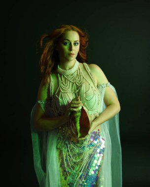 Peri masalı kostümü giymiş kızıl saçlı bir deniz kızı perisinin fantezi stüdyosu portresi. Klasik poz, deniz kabuğunu havada süzülen kumaş ve ruhsal kasvetli stüdyo ışıklandırmasıyla tutuyordu.