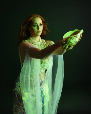 Peri masalı kostümü giymiş kızıl saçlı bir deniz kızı perisinin fantezi stüdyosu portresi. Klasik poz, deniz kabuğunu havada süzülen kumaş ve ruhsal kasvetli stüdyo ışıklandırmasıyla tutuyordu.