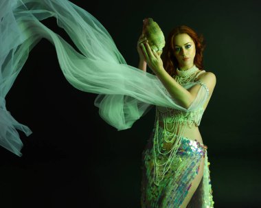 Peri masalı kostümü giymiş kızıl saçlı bir deniz kızı perisinin fantezi stüdyosu portresi. Klasik poz, deniz kabuğunu havada süzülen kumaş ve ruhsal kasvetli stüdyo ışıklandırmasıyla tutuyordu.