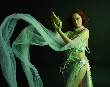 Peri masalı kostümü giymiş kızıl saçlı bir deniz kızı perisinin fantezi stüdyosu portresi. Klasik poz, deniz kabuğunu havada süzülen kumaş ve ruhsal kasvetli stüdyo ışıklandırmasıyla tutuyordu.