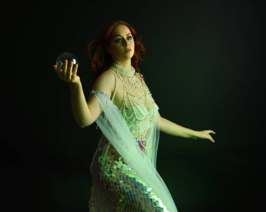 Fantezi stüdyo portresi, beyaz inci elbise giyen kızıl saçlı bir deniz kızı peri modeli, peri masalı kostümü. Akan kumaşlı, ruhsal kasvetli stüdyo ışıklandırmalı klasik poz.