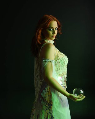 Fantezi stüdyo portresi, beyaz inci elbise giyen kızıl saçlı bir deniz kızı peri modeli, peri masalı kostümü. Akan kumaşlı, ruhsal kasvetli stüdyo ışıklandırmalı klasik poz.