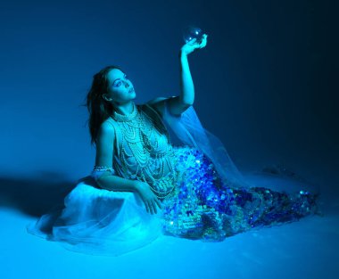 Fantezi stüdyo portresi, beyaz inci elbise giyen kızıl saçlı bir deniz kızı peri modeli, peri masalı kostümü. Klasik oturma pozu, akıcı kumaş, ruhani renkli neon ışık stüdyo arka planında.