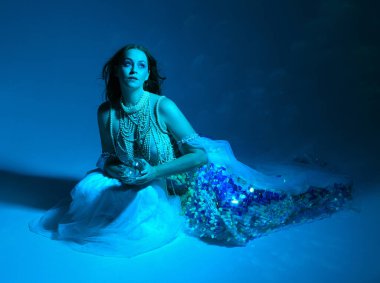 Fantezi stüdyo portresi, beyaz inci elbise giyen kızıl saçlı bir deniz kızı peri modeli, peri masalı kostümü. Klasik oturma pozu, akıcı kumaş, ruhani renkli neon ışık stüdyo arka planında.