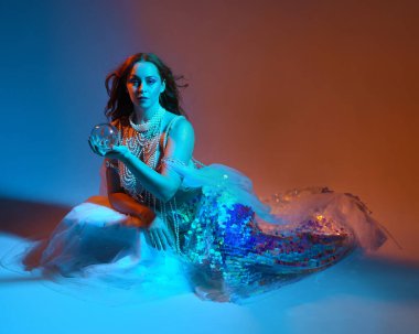 Fantezi stüdyo portresi, beyaz inci elbise giyen kızıl saçlı bir deniz kızı peri modeli, peri masalı kostümü. Klasik oturma pozu, akıcı kumaş, ruhani renkli neon ışık stüdyo arka planında.