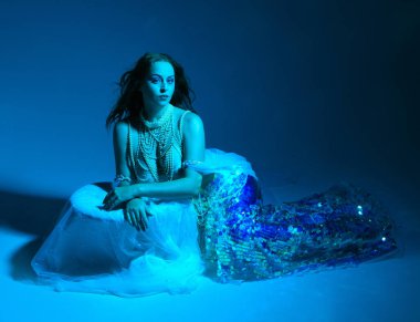 Fantezi stüdyo portresi, beyaz inci elbise giyen kızıl saçlı bir deniz kızı peri modeli, peri masalı kostümü. Klasik oturma pozu, akıcı kumaş, ruhani renkli neon ışık stüdyo arka planında.