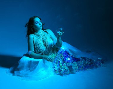 Fantezi stüdyo portresi, beyaz inci elbise giyen kızıl saçlı bir deniz kızı peri modeli, peri masalı kostümü. Klasik oturma pozu, akıcı kumaş, ruhani renkli neon ışık stüdyo arka planında.