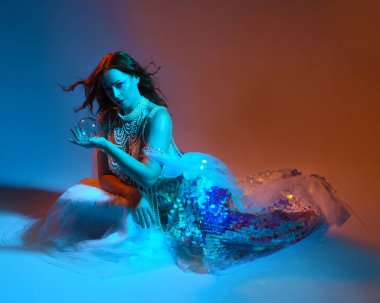 Fantezi stüdyo portresi, beyaz inci elbise giyen kızıl saçlı bir deniz kızı peri modeli, peri masalı kostümü. Klasik oturma pozu, akıcı kumaş, ruhani renkli neon ışık stüdyo arka planında.