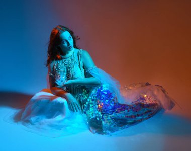 Fantezi stüdyo portresi, beyaz inci elbise giyen kızıl saçlı bir deniz kızı peri modeli, peri masalı kostümü. Klasik oturma pozu, akıcı kumaş, ruhani renkli neon ışık stüdyo arka planında.