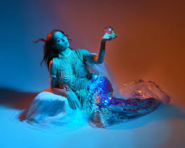 Fantezi stüdyo portresi, beyaz inci elbise giyen kızıl saçlı bir deniz kızı peri modeli, peri masalı kostümü. Klasik oturma pozu, akıcı kumaş, ruhani renkli neon ışık stüdyo arka planında.