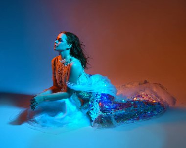 Fantezi stüdyo portresi, beyaz inci elbise giyen kızıl saçlı bir deniz kızı peri modeli, peri masalı kostümü. Klasik oturma pozu, akıcı kumaş, ruhani renkli neon ışık stüdyo arka planında.