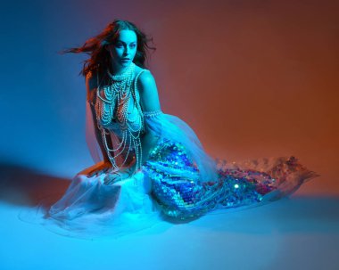 Fantezi stüdyo portresi, beyaz inci elbise giyen kızıl saçlı bir deniz kızı peri modeli, peri masalı kostümü. Klasik oturma pozu, akıcı kumaş, ruhani renkli neon ışık stüdyo arka planında.