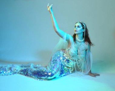 Fantezi stüdyo portresi, beyaz inci elbise giyen kızıl saçlı bir deniz kızı peri modeli, peri masalı kostümü. Klasik oturma pozisyonu, ruhsal kasvetli mavi ışıklandırma stüdyo arka planında su altındaymış gibi.