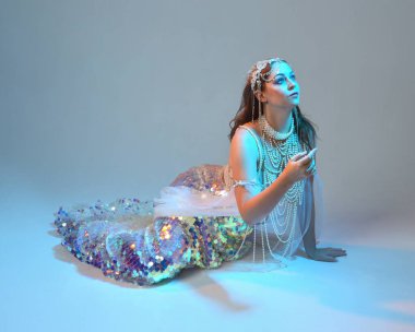 Fantezi stüdyo portresi, beyaz inci elbise giyen kızıl saçlı bir deniz kızı peri modeli, peri masalı kostümü. Klasik oturma pozisyonu, ruhsal kasvetli mavi ışıklandırma stüdyo arka planında su altındaymış gibi.