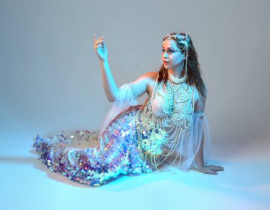 Fantezi stüdyo portresi, beyaz inci elbise giyen kızıl saçlı bir deniz kızı peri modeli, peri masalı kostümü. Klasik oturma pozisyonu, ruhsal kasvetli mavi ışıklandırma stüdyo arka planında su altındaymış gibi.