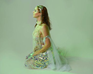 Fantezi stüdyo portresi, beyaz inci elbise giyen kızıl saçlı bir deniz kızı peri modeli, peri masalı kostümü. Klasik oturma pozisyonu, ruhsal kasvetli mavi ışıklandırma stüdyo arka planında su altındaymış gibi.