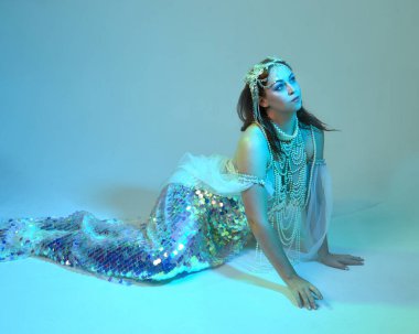 Fantezi stüdyo portresi, beyaz inci elbise giyen kızıl saçlı bir deniz kızı peri modeli, peri masalı kostümü. Klasik oturma pozisyonu, ruhsal kasvetli mavi ışıklandırma stüdyo arka planında su altındaymış gibi.