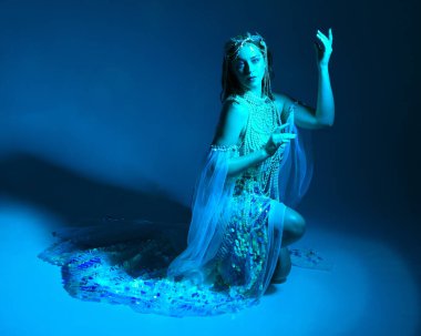 Fantezi stüdyo portresi, beyaz inci elbise giyen kızıl saçlı bir deniz kızı peri modeli, peri masalı kostümü. Klasik oturma pozisyonu, ruhsal kasvetli mavi ışıklandırma stüdyo arka planında su altındaymış gibi.