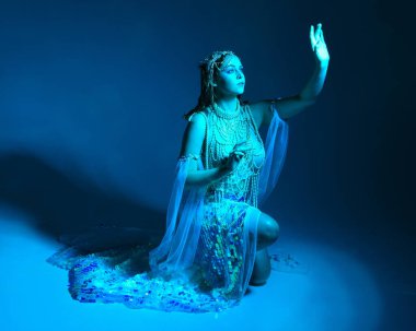 Fantezi stüdyo portresi, beyaz inci elbise giyen kızıl saçlı bir deniz kızı peri modeli, peri masalı kostümü. Klasik oturma pozisyonu, ruhsal kasvetli mavi ışıklandırma stüdyo arka planında su altındaymış gibi.