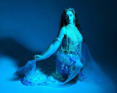 Fantezi stüdyo portresi, beyaz inci elbise giyen kızıl saçlı bir deniz kızı peri modeli, peri masalı kostümü. Klasik oturma pozisyonu, ruhsal kasvetli mavi ışıklandırma stüdyo arka planında su altındaymış gibi.