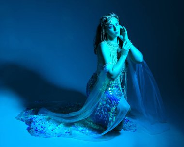 Fantezi stüdyo portresi, beyaz inci elbise giyen kızıl saçlı bir deniz kızı peri modeli, peri masalı kostümü. Klasik oturma pozisyonu, ruhsal kasvetli mavi ışıklandırma stüdyo arka planında su altındaymış gibi.