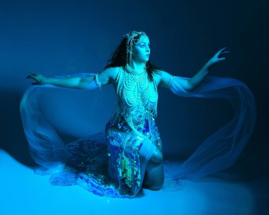 Fantezi stüdyo portresi, beyaz inci elbise giyen kızıl saçlı bir deniz kızı peri modeli, peri masalı kostümü. Klasik oturma pozisyonu, ruhsal kasvetli mavi ışıklandırma stüdyo arka planında su altındaymış gibi.