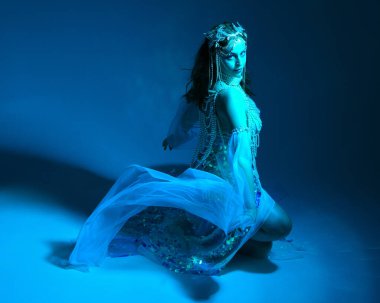 Fantezi stüdyo portresi, beyaz inci elbise giyen kızıl saçlı bir deniz kızı peri modeli, peri masalı kostümü. Klasik oturma pozisyonu, ruhsal kasvetli mavi ışıklandırma stüdyo arka planında su altındaymış gibi.