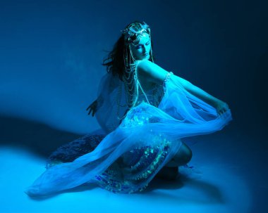 Fantezi stüdyo portresi, beyaz inci elbise giyen kızıl saçlı bir deniz kızı peri modeli, peri masalı kostümü. Klasik oturma pozisyonu, ruhsal kasvetli mavi ışıklandırma stüdyo arka planında su altındaymış gibi.