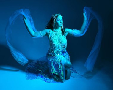 Fantezi stüdyo portresi, beyaz inci elbise giyen kızıl saçlı bir deniz kızı peri modeli, peri masalı kostümü. Klasik oturma pozisyonu, ruhsal kasvetli mavi ışıklandırma stüdyo arka planında su altındaymış gibi.