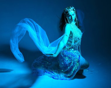 Fantezi stüdyo portresi, beyaz inci elbise giyen kızıl saçlı bir deniz kızı peri modeli, peri masalı kostümü. Klasik oturma pozisyonu, ruhsal kasvetli mavi ışıklandırma stüdyo arka planında su altındaymış gibi.