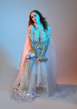 Peri masalı kostümü giymiş kızıl saçlı bir deniz kızı perisinin fantezi stüdyosu portresi. Klasik oturma pozu akışkan kumaş ve ruhani stüdyo ışıklandırması, izole edilmiş arka plan.