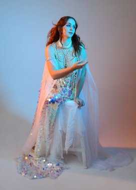 Peri masalı kostümü giymiş kızıl saçlı bir deniz kızı perisinin fantezi stüdyosu portresi. Klasik oturma pozu akışkan kumaş ve ruhani stüdyo ışıklandırması, izole edilmiş arka plan.