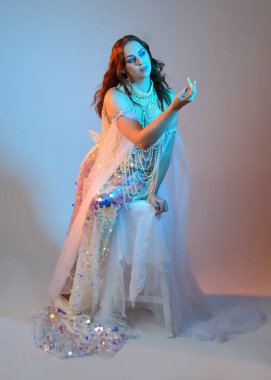 Peri masalı kostümü giymiş kızıl saçlı bir deniz kızı perisinin fantezi stüdyosu portresi. Klasik oturma pozu akışkan kumaş ve ruhani stüdyo ışıklandırması, izole edilmiş arka plan.