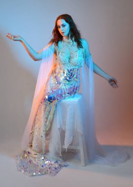 Peri masalı kostümü giymiş kızıl saçlı bir deniz kızı perisinin fantezi stüdyosu portresi. Klasik oturma pozu akışkan kumaş ve ruhani stüdyo ışıklandırması, izole edilmiş arka plan.