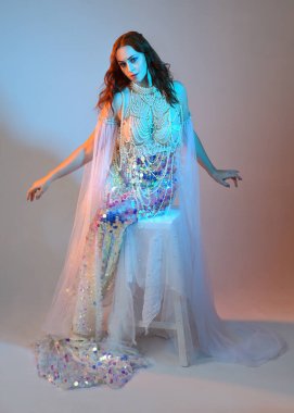 Peri masalı kostümü giymiş kızıl saçlı bir deniz kızı perisinin fantezi stüdyosu portresi. Klasik oturma pozu akışkan kumaş ve ruhani stüdyo ışıklandırması, izole edilmiş arka plan.