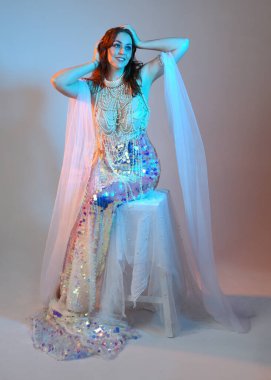 Peri masalı kostümü giymiş kızıl saçlı bir deniz kızı perisinin fantezi stüdyosu portresi. Klasik oturma pozu akışkan kumaş ve ruhani stüdyo ışıklandırması, izole edilmiş arka plan.