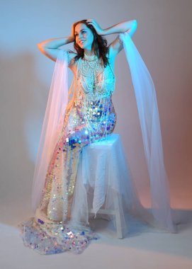 Peri masalı kostümü giymiş kızıl saçlı bir deniz kızı perisinin fantezi stüdyosu portresi. Klasik oturma pozu akışkan kumaş ve ruhani stüdyo ışıklandırması, izole edilmiş arka plan.