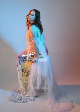 Peri masalı kostümü giymiş kızıl saçlı bir deniz kızı perisinin fantezi stüdyosu portresi. Klasik oturma pozu akışkan kumaş ve ruhani stüdyo ışıklandırması, izole edilmiş arka plan.