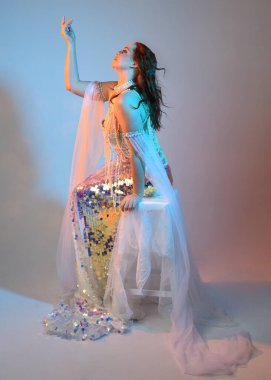 Peri masalı kostümü giymiş kızıl saçlı bir deniz kızı perisinin fantezi stüdyosu portresi. Klasik oturma pozu akışkan kumaş ve ruhani stüdyo ışıklandırması, izole edilmiş arka plan.