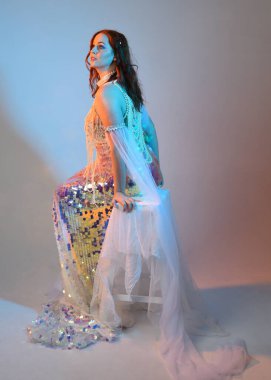 Peri masalı kostümü giymiş kızıl saçlı bir deniz kızı perisinin fantezi stüdyosu portresi. Klasik oturma pozu akışkan kumaş ve ruhani stüdyo ışıklandırması, izole edilmiş arka plan.