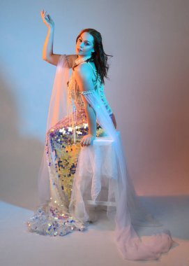 Peri masalı kostümü giymiş kızıl saçlı bir deniz kızı perisinin fantezi stüdyosu portresi. Klasik oturma pozu akışkan kumaş ve ruhani stüdyo ışıklandırması, izole edilmiş arka plan.