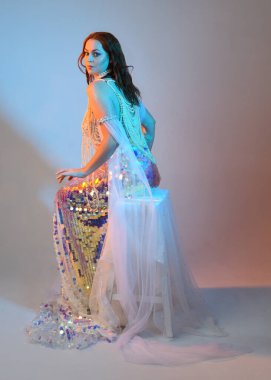 Peri masalı kostümü giymiş kızıl saçlı bir deniz kızı perisinin fantezi stüdyosu portresi. Klasik oturma pozu akışkan kumaş ve ruhani stüdyo ışıklandırması, izole edilmiş arka plan.