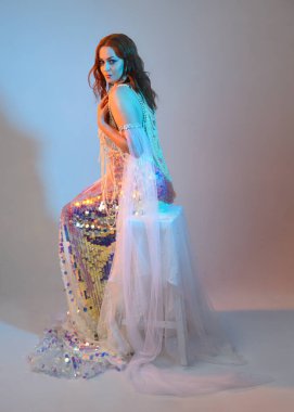 Peri masalı kostümü giymiş kızıl saçlı bir deniz kızı perisinin fantezi stüdyosu portresi. Klasik oturma pozu akışkan kumaş ve ruhani stüdyo ışıklandırması, izole edilmiş arka plan.