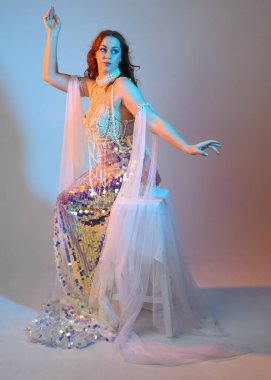 Peri masalı kostümü giymiş kızıl saçlı bir deniz kızı perisinin fantezi stüdyosu portresi. Klasik oturma pozu akışkan kumaş ve ruhani stüdyo ışıklandırması, izole edilmiş arka plan.