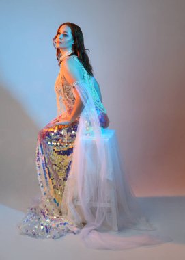 Peri masalı kostümü giymiş kızıl saçlı bir deniz kızı perisinin fantezi stüdyosu portresi. Klasik oturma pozu akışkan kumaş ve ruhani stüdyo ışıklandırması, izole edilmiş arka plan.