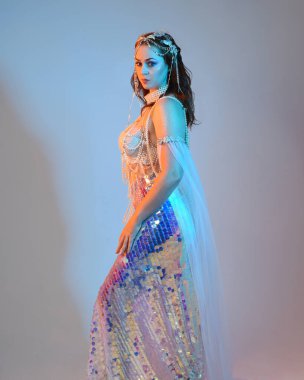 Peri masalı kostümü giymiş kızıl saçlı bir deniz kızı perisinin fantezi stüdyosu portresi. Akan kumaş ve ruhani stüdyo ışıklandırmasıyla klasik yürüyüş pozu, izole edilmiş arka plan.