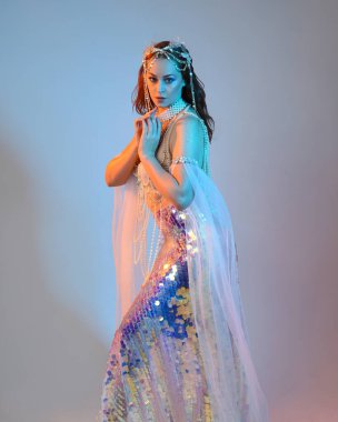 Peri masalı kostümü giymiş kızıl saçlı bir deniz kızı perisinin fantezi stüdyosu portresi. Akan kumaş ve ruhani stüdyo ışıklandırmasıyla klasik yürüyüş pozu, izole edilmiş arka plan.