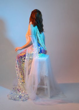 Peri masalı kostümü giymiş kızıl saçlı bir deniz kızı perisinin fantezi stüdyosu portresi. Klasik oturma pozu akışkan kumaş ve ruhani stüdyo ışıklandırması, izole edilmiş arka plan.