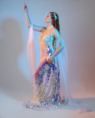 Peri masalı kostümü giymiş kızıl saçlı bir deniz kızı perisinin fantezi stüdyosu portresi. Akan kumaş ve ruhani stüdyo ışıklandırmasıyla klasik yürüyüş pozu, izole edilmiş arka plan.